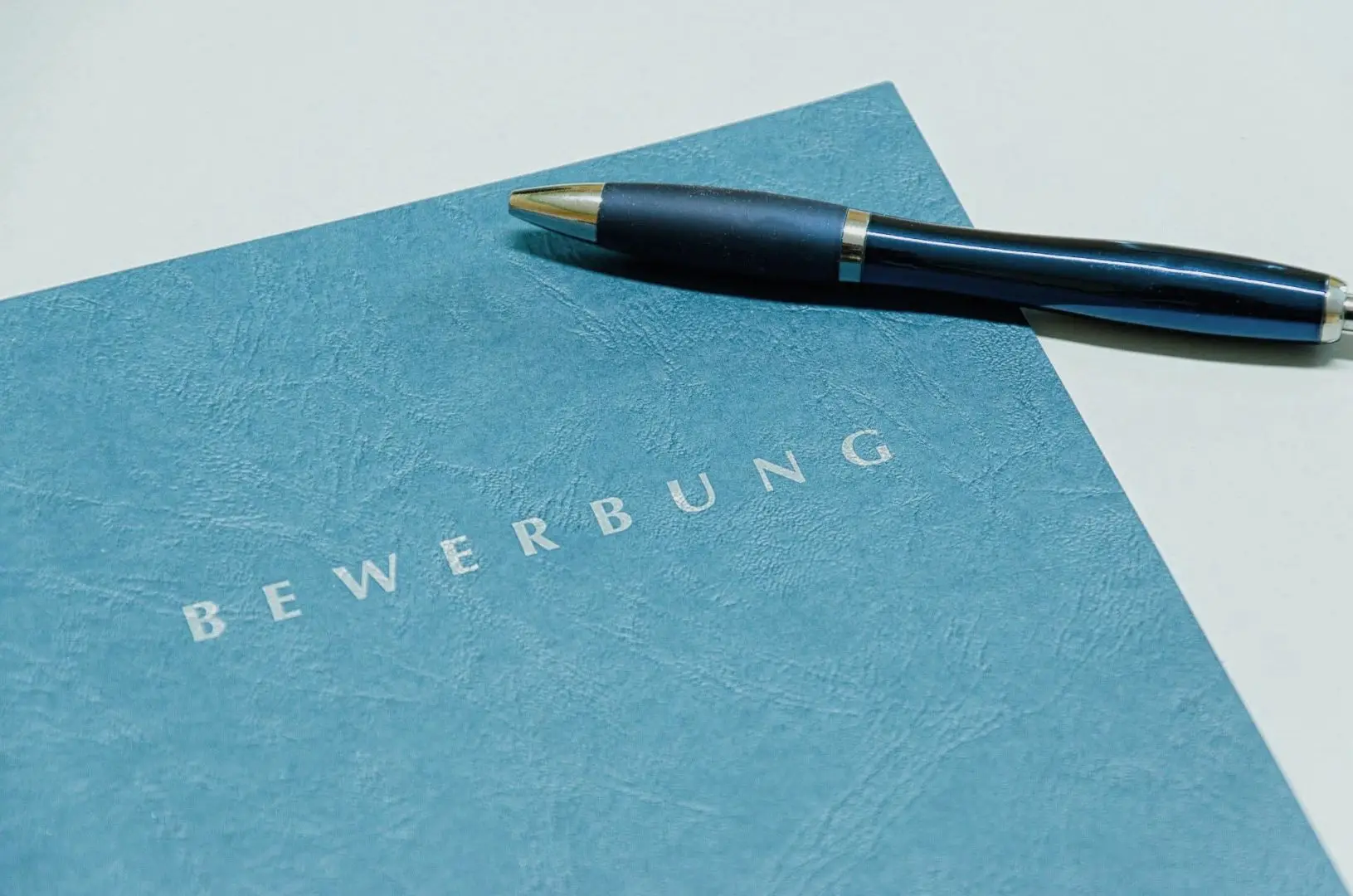 Stilvoller Stift auf einem blauen Ordner mit Aufschrift 'BEWERBUNG', symbolisiert professionelles AVGS-Coaching in ganz Deutschland.