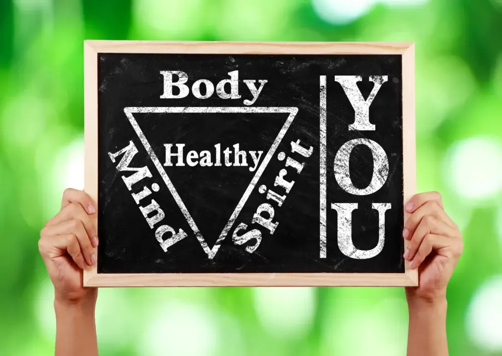 Hände halten eine Tafel mit der Aufschrift 'Body, Mind, Spirit – Healthy You', symbolisch für ganzheitliches Wohlbefinden und Selbstfürsorge durch AVGS-Coaching.