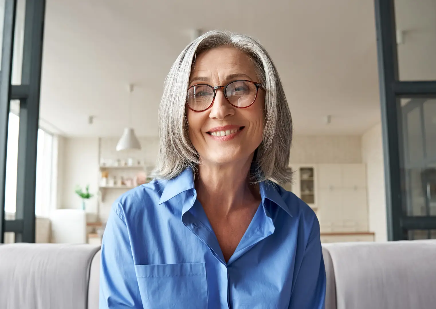 Eine ältere Frau mit grauen Haaren und brauner Brille, die lächelnd in die Kamera schaut und gerade ein Onlinecoaching absolviert.
