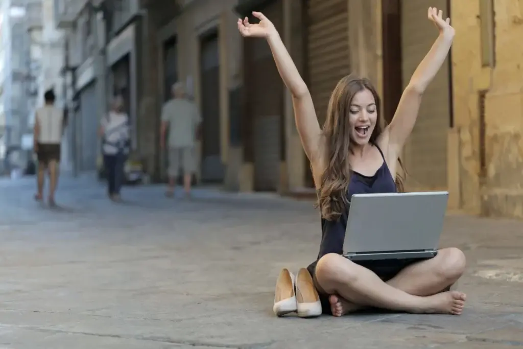 Freudige junge Frau sitzt auf dem Boden in einer urbanen Straßenumgebung und jubelt mit erhobenen Armen beim Blick auf ihren Laptop, was den Erfolg und das Empowerment durch Coaching symbolisiert.