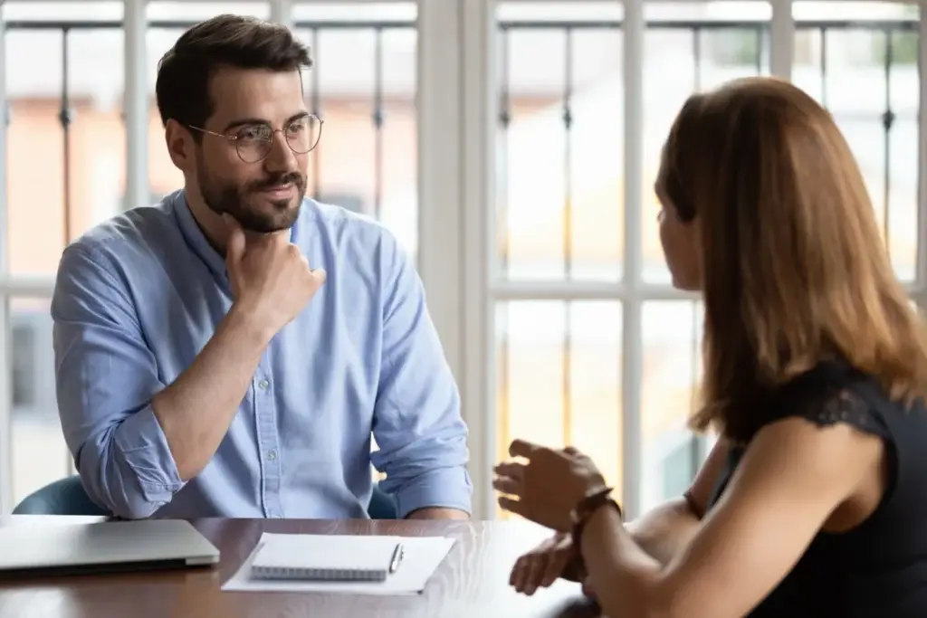 Selbstbewusster junger Geschäftsmann mit Brille und blauem Hemd führt ein Gespräch mit einer Kollegin in einem hellen Büro zum Thema Jobcoaching.